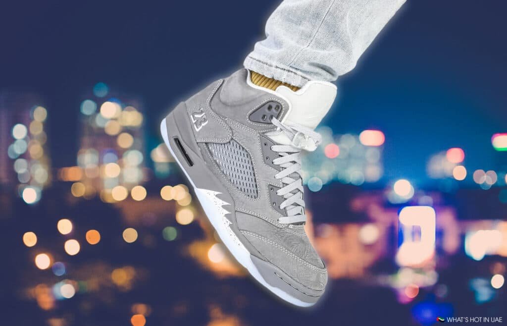 A gray high-top sneaker with white accents and lace details, worn with light blue jeans, set against a blurred city lights background at night.