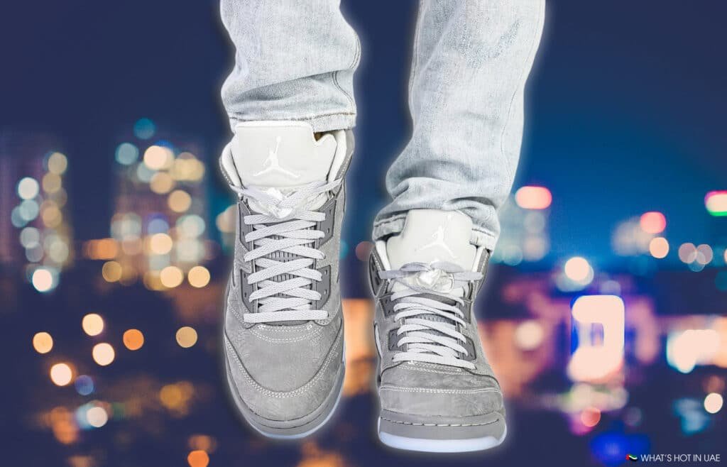 Close-up of a person wearing light gray sneakers with a white logo, and light blue jeans, standing against a blurred cityscape with colorful bokeh lights.
