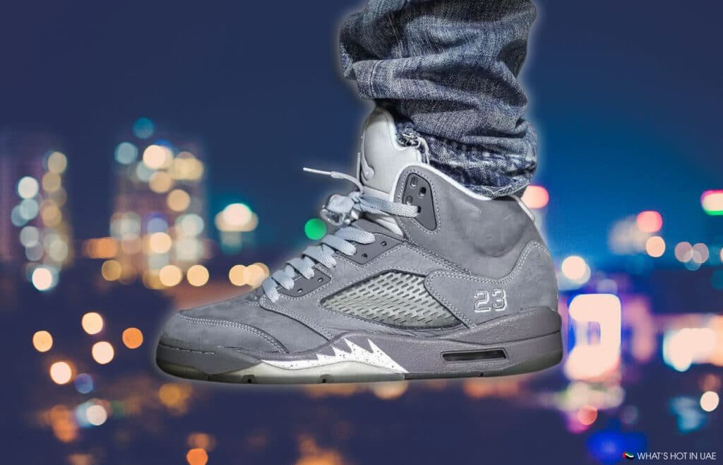A close-up of a Air Jordan 5 Wolf Grey sneaker on a person's foot against a blurred cityscape at night. The shoe features a number '23' embroidered on the side, with detailed laces and a mesh panel.