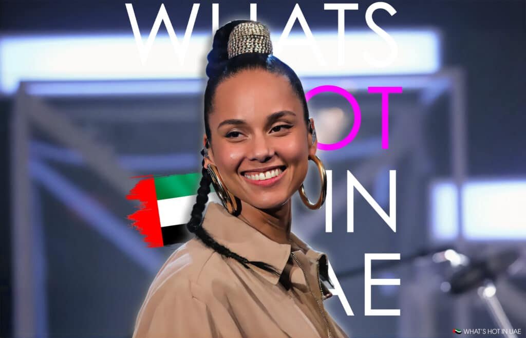 A person with a stylish braided bun hairstyle and large hoop earrings smiles against a blurred background featuring text and a UAE flag, with "What's Hot in UAE" written on it.