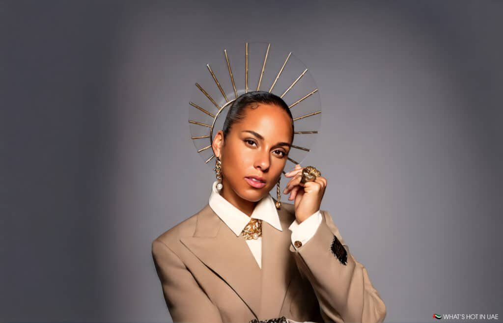 Alicia Keys at Saadiyat Nights wearing a tan blazer and white collared shirt, adorned with a gold statement necklace and earrings, poses against a neutral background with a decorative golden headpiece resembling a sunburst.