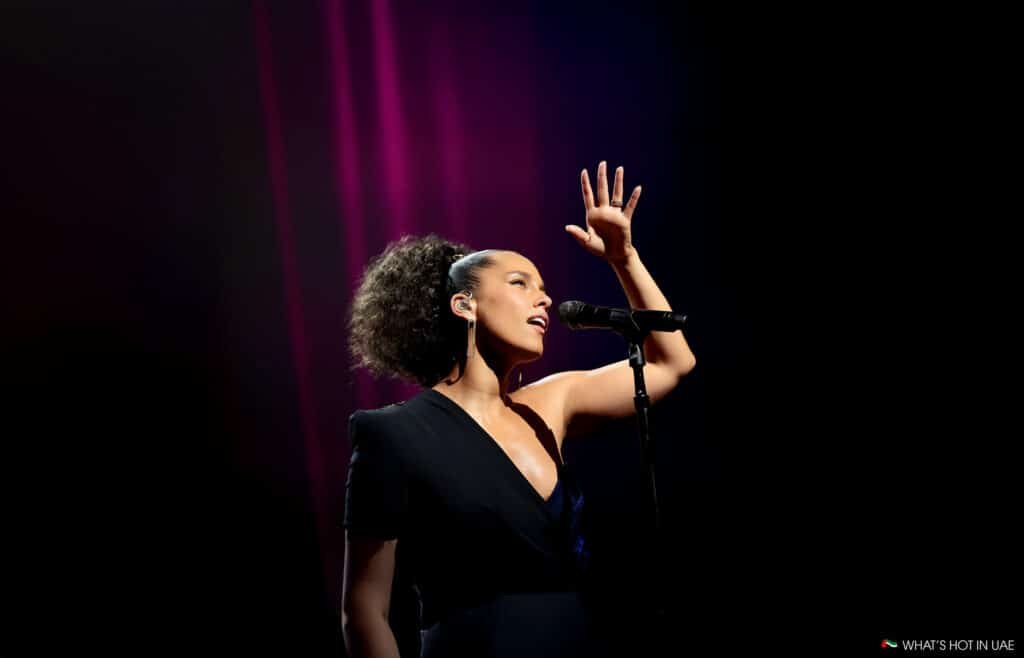 Person singing into a microphone on stage, with dramatic pink lighting in the background, wearing a dark, one-shoulder outfit and earrings.