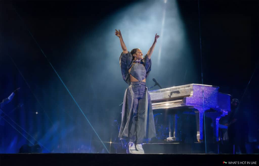 Alicia Keys at Saadiyat Nights stands on stage with arms raised, in front of a sparkling grand piano, under a spotlight during a performance.