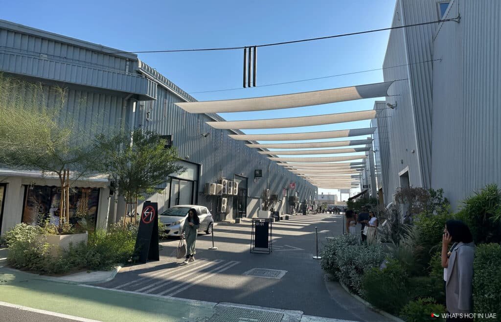 Alserkal Avenue with modern industrial buildings featuring metal facades, outdoor seating, and people walking under shade structures in a lively urban area.