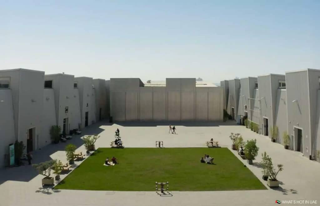 An open Alserkal Avenue with a green grassy area surrounded by industrial-style buildings on a clear day. People are sitting on the grass and benches, and some are walking along the perimeter.