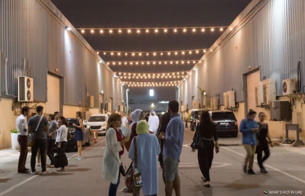 A group of people socializing in a well-lit Alserkal Avenue at night, lined with industrial buildings and decorated with string lights overhead.