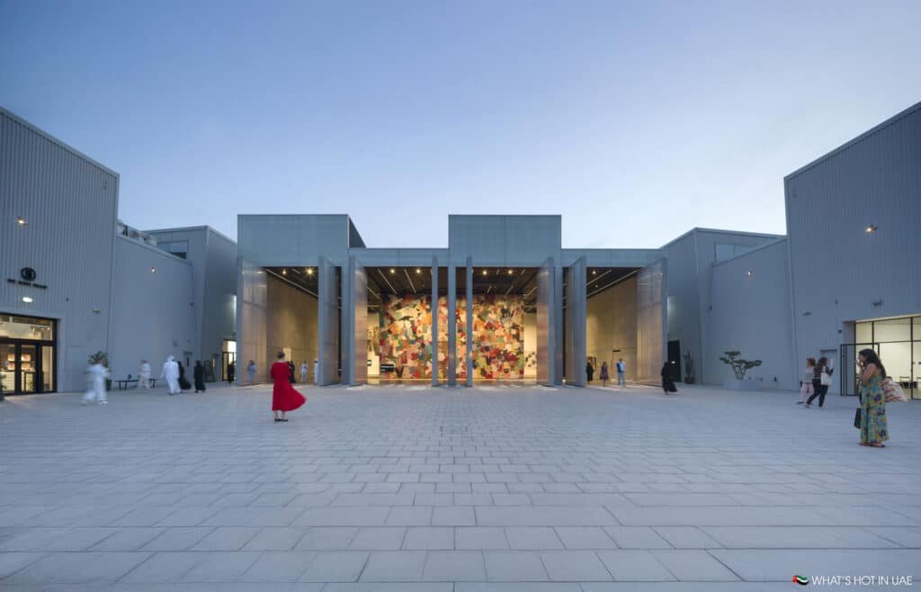 A modern building with a spacious courtyard, featuring a colorful mural on the interior wall visible through open doors, surrounded by people walking and standing on a tiled plaza at dusk.