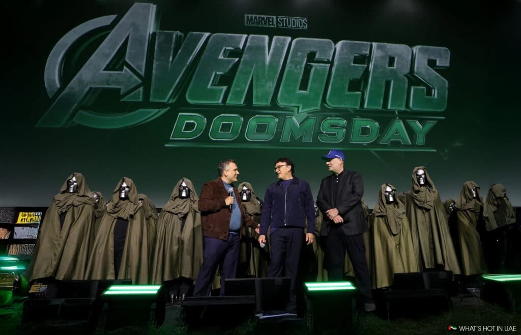 Three men standing in front of a "Avengers: Doomsday " sign with multiple cloaked figures behind them.