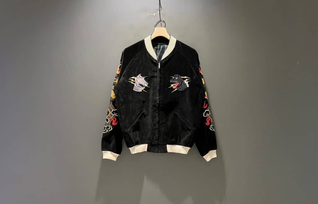 A black Beams Japan x Tailor Toyo jacket with colorful embroidered designs on the front and sleeves hangs against a gray background.