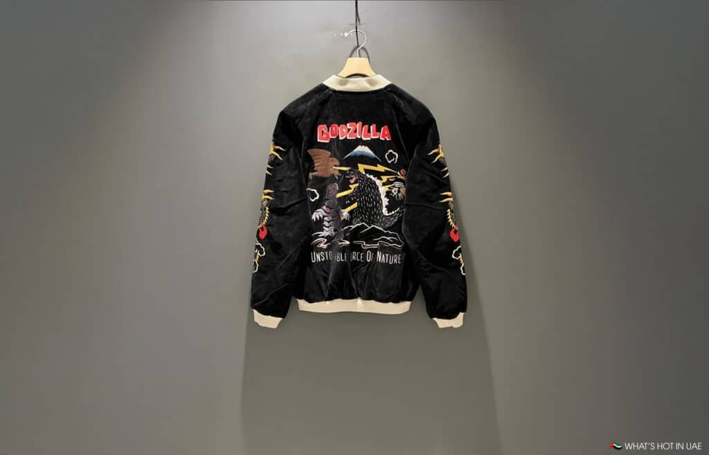 A black bomber jacket with a colorful Godzilla design and decorative text hangs on a wall.