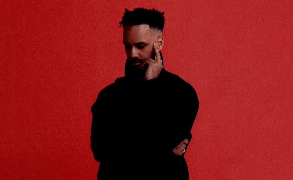 A person in a dark outfit stands against a bright red background, with one hand touching their face in a contemplative pose.