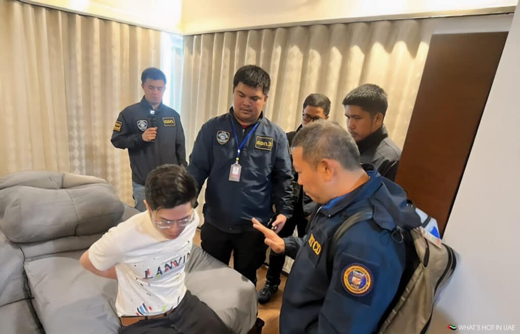 A group of uniformed officers stands around a Chinese crypto scammers seated man with his hands restrained behind him in a room.