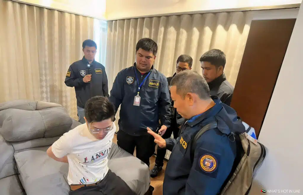 A group of uniformed officers stands around a Chinese crypto scammers seated man with his hands restrained behind him in a room.
