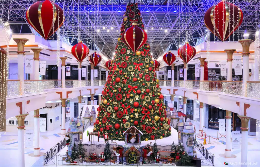 A large Christmas tree decorated with red bows, ornaments, and lights stands in a brightly lit mall atrium, surrounded by oversized red and gold ornaments hanging from the ceiling.