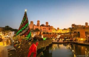 A large Christmas tree decorated with lights and ornaments stands next to a waterway, with traditional Arabian style buildings and palm trees in the background at dusk.