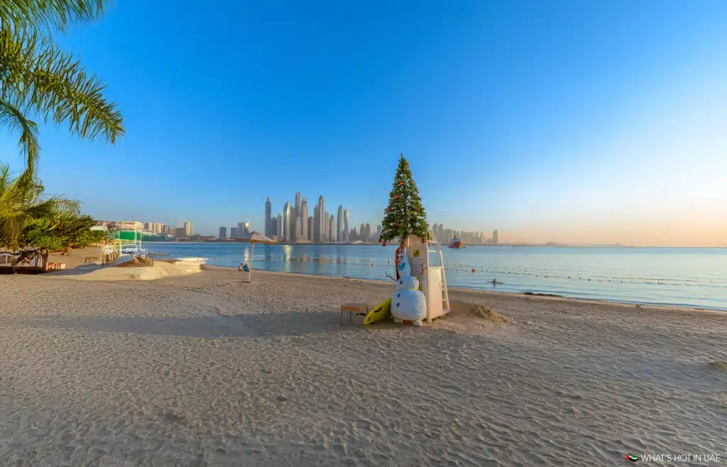A beach with a decorated Christmas tree and snowman figure on a lifeguard stand, with a view of a city's skyline in the background under a clear blue sky.