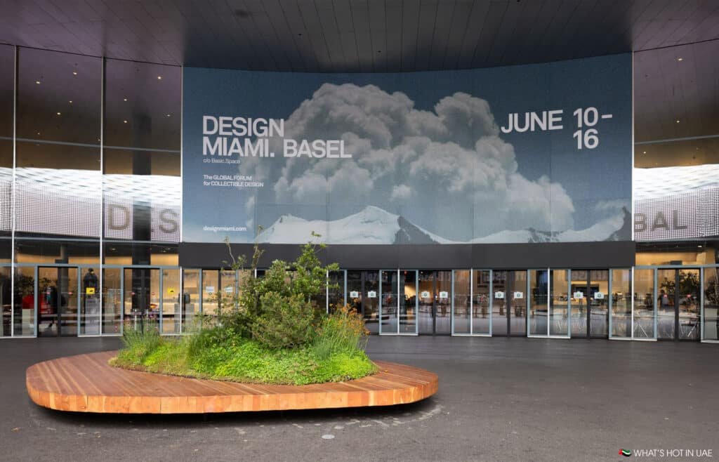 The entrance of a building with a large billboard advertising "Design Miami. Basel" with the dates June 10-16, featuring an image of mountains and clouds. In front, there is a wooden platform with plants and greenery.