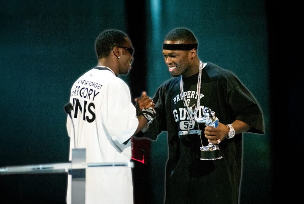 P Diddy & 50 Cent on stage, one holding a silver award statue while they shake hands and smile at each other. One wears a white jersey with text, and the other wears a black T-shirt with a headband.