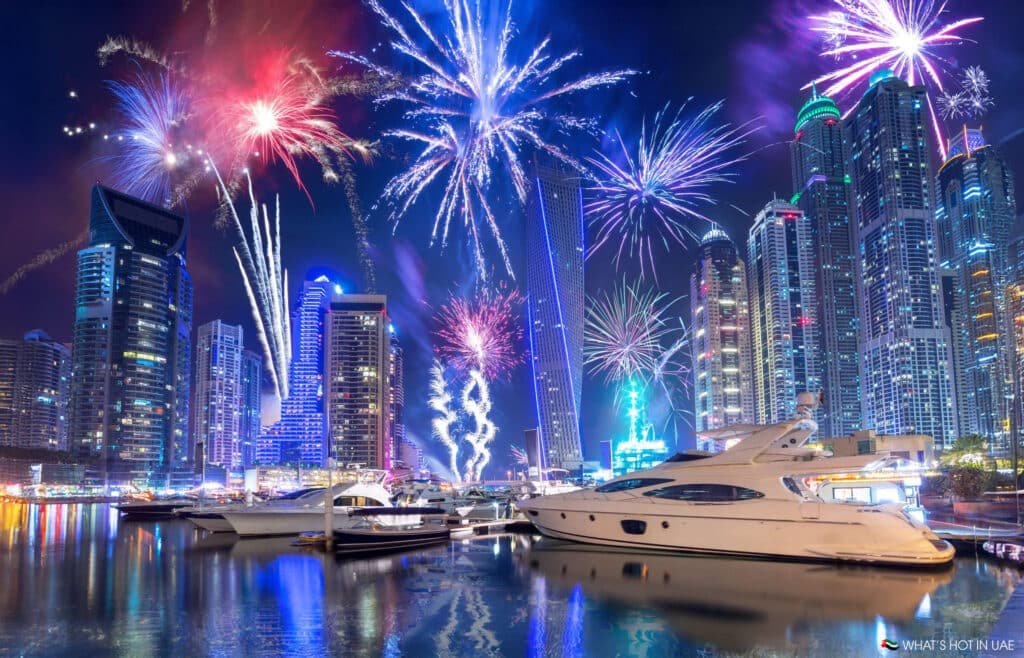 NYE 2026 Fireworks illuminate a marina with several yachts, surrounded by a skyline of lit-up skyscrapers at night.