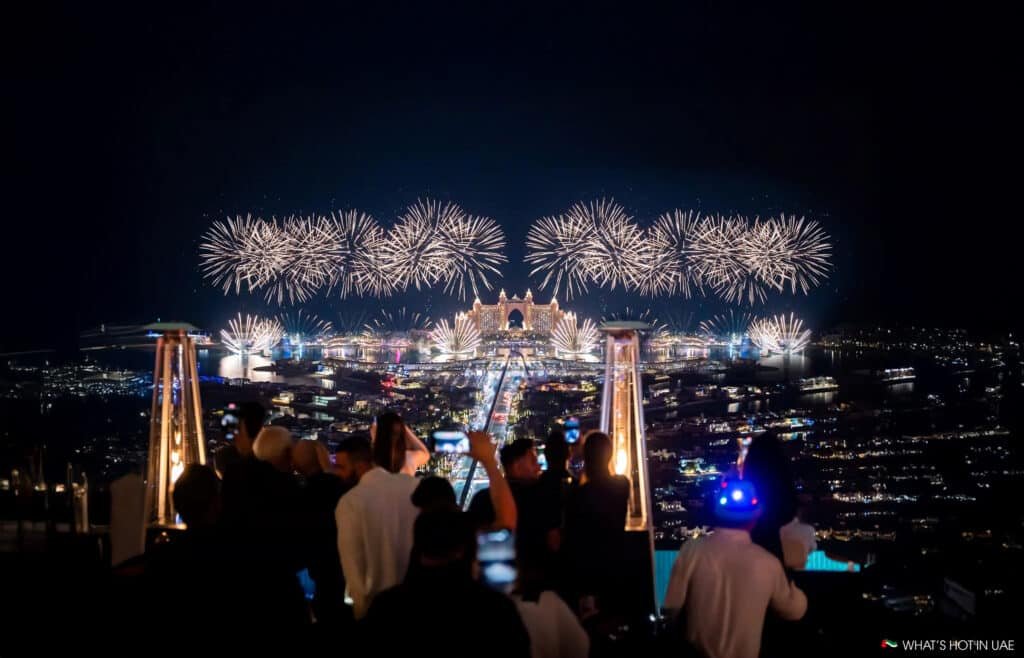 People watch a large NYE 2026 fireworks display illuminating a cityscape at night.
