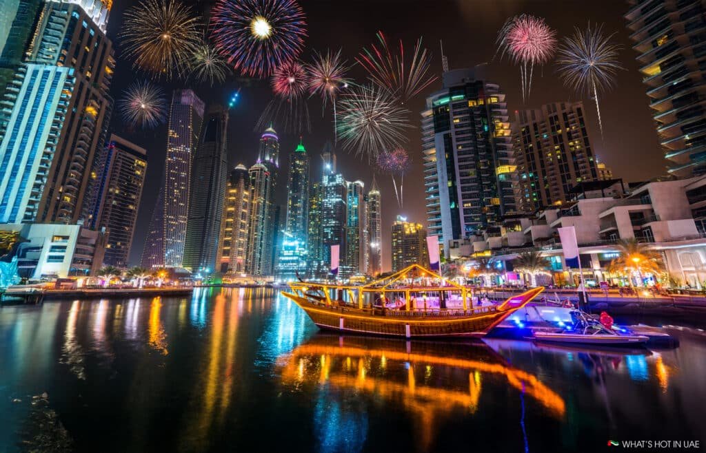 A brightly lit boat on a reflective waterway with fireworks above a city skyline at night.