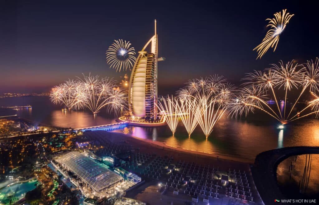 Fireworks illuminate the night sky around a sail-shaped skyscraper on a coastline with a lively waterfront scene.
