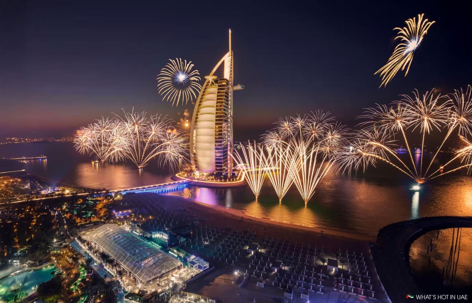 Fireworks illuminate the night sky around a sail-shaped skyscraper on a coastline with a lively waterfront scene.