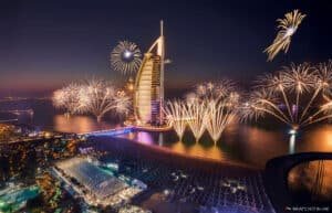 Fireworks illuminate the night sky around a sail-shaped skyscraper on a coastline with a lively waterfront scene.