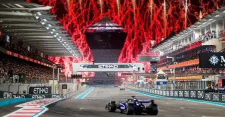 A Formula 1 car racing down the Yas Marina Circuit during a nighttime event with red fireworks lighting up the sky in the background, surrounded by spectators in the grandstands.