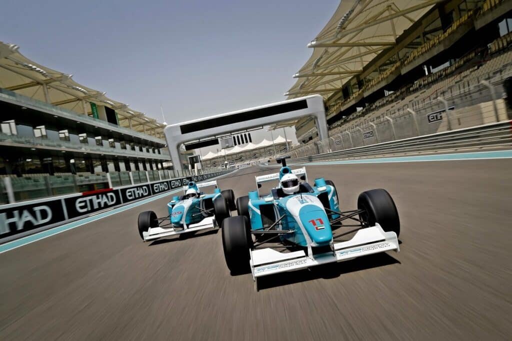 Two racing cars with blue and white livery driving side by side on a track under a large gantry, with grandstands in the background.