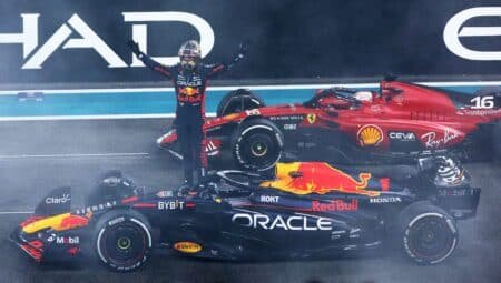 A Formula 1 driver in a Red Bull racing suit stands beside a Red Bull race car during a pause in an event, with a Ferrari race car in the background, and track signage visible above.