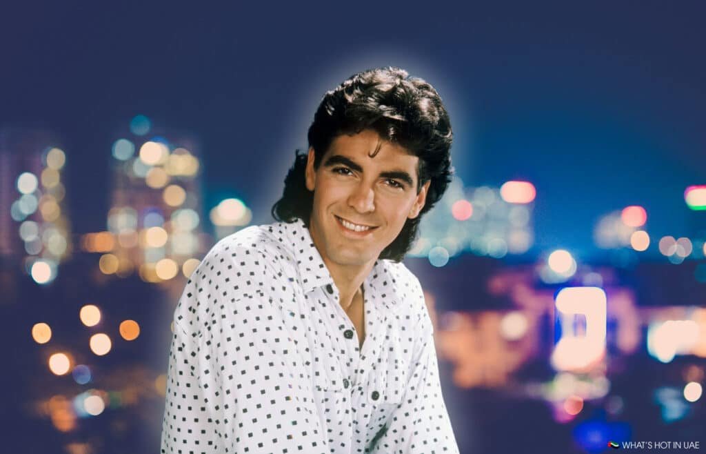 George Clooney with curly hair wearing a white shirt with black dots standing against a blurry cityscape at night.