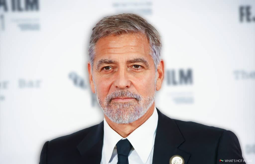 A man with short gray hair and a beard is wearing a black suit and tie, standing against a blurred backdrop with logos in the background.