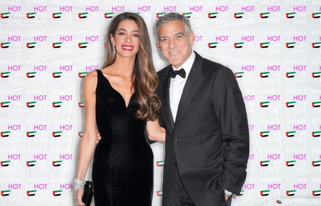 A well-dressed George and Amal Clooney posing in front of a backdrop with 'WHAT'S HOT IN UAE' and UAE flags repeatedly printed on it; the woman wears a black dress, and the man wears a black tuxedo.