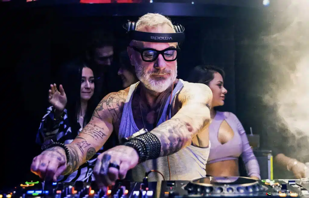 A DJ at Nikki Beach Dubai wearing glasses and headphones, operating a sound mixer in a nightclub setting, surrounded by dancing people.