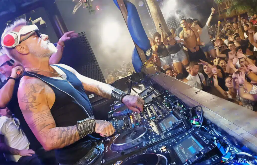 A DJ at Nikki Beach Dubai with tattoos and wearing sunglasses performs at a lively club, playing music on professional equipment while a large crowd enjoys the party atmosphere.