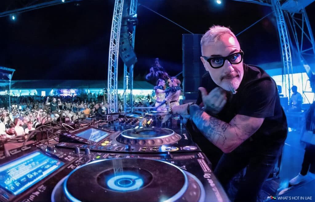 A DJ with a beard and tattoos playing music at an outdoor event, with a large crowd and intricate lighting setup visible in the background.