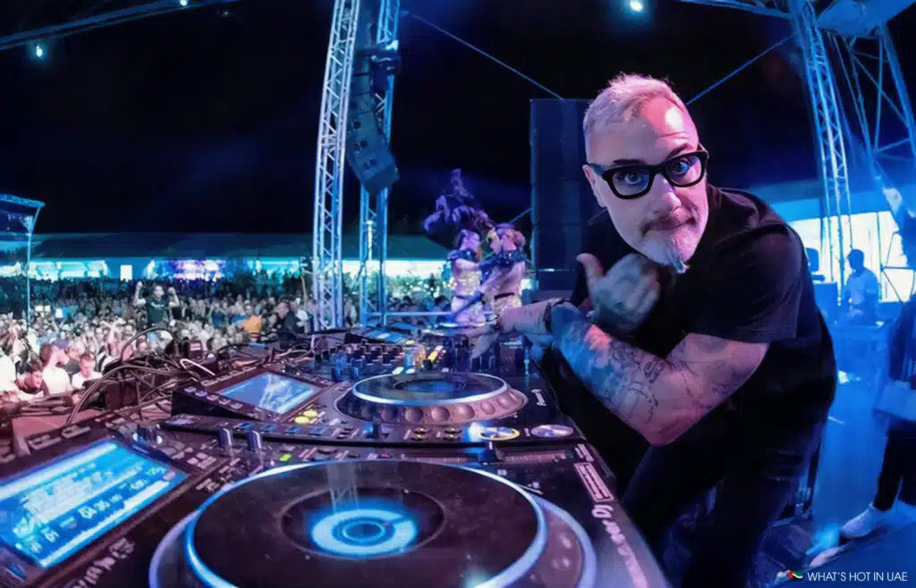 A DJ with a beard and tattoos playing music at an outdoor event, with a large crowd and intricate lighting setup visible in the background.