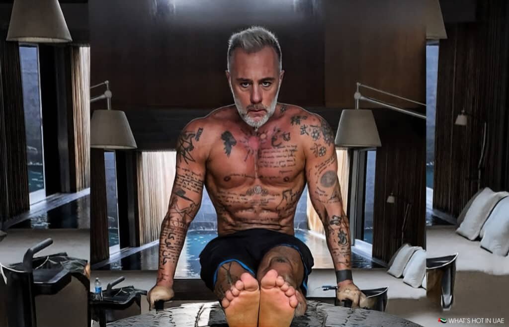 A tattooed man with gray hair and a beard performs a strength exercise indoors, balancing his body on parallel bars with his legs extended forward. Lamps and a pool are visible in the background.