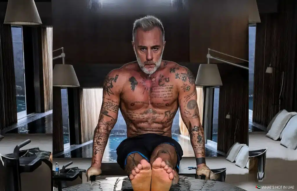 A tattooed man with gray hair and a beard performs a strength exercise indoors, balancing his body on parallel bars with his legs extended forward. Lamps and a pool are visible in the background.