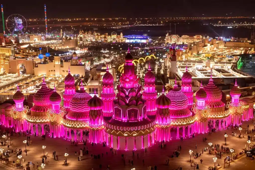 A vibrant nighttime scene of Global Village, showcasing a large, illuminated building with domes and a lively background of rides and lights.