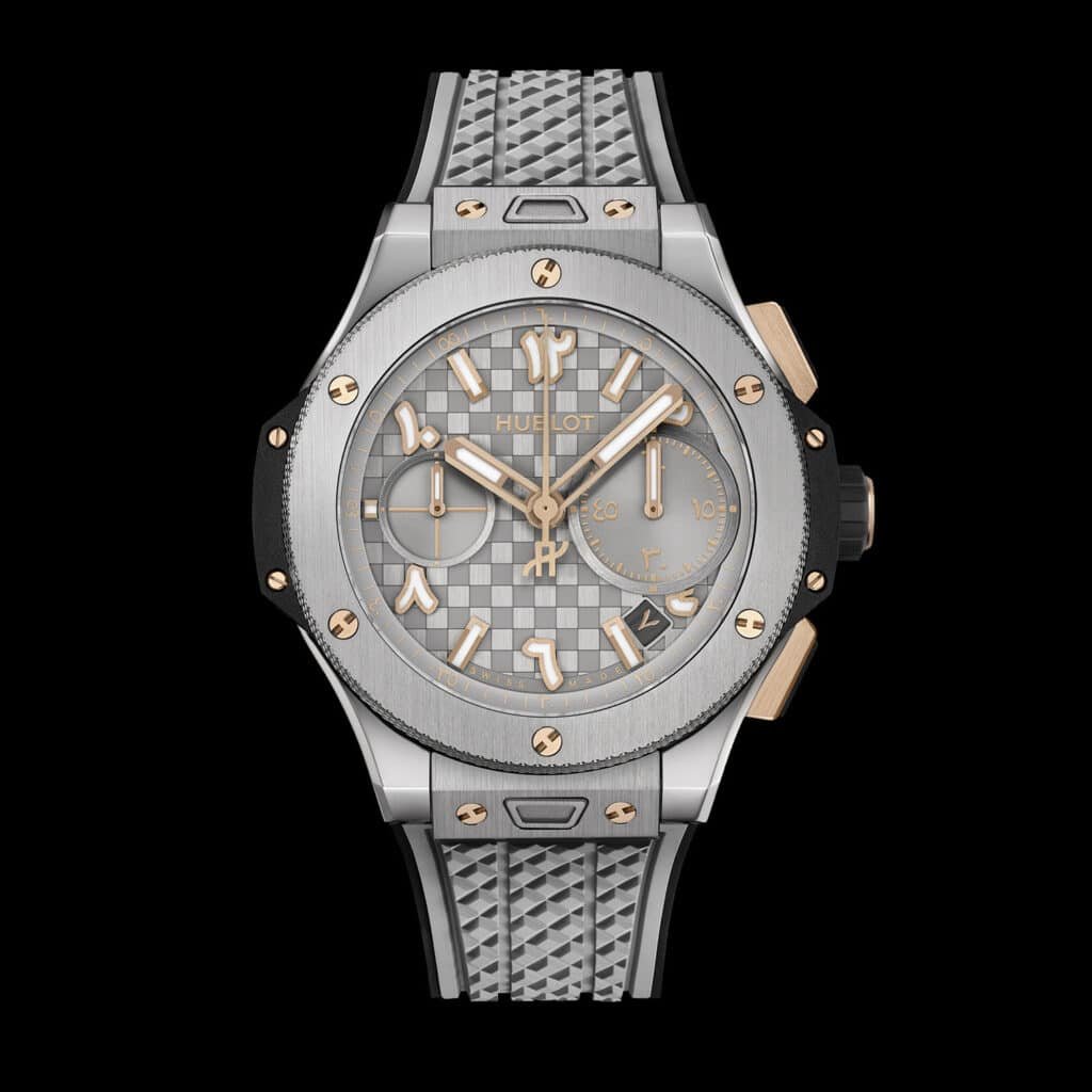 A luxury wristwatch with a silver and rose gold-toned bezel and a patterned grey face. It features Arabic numeral hour markers, three sub-dials, and a date display. The strap is grey with a textured design.