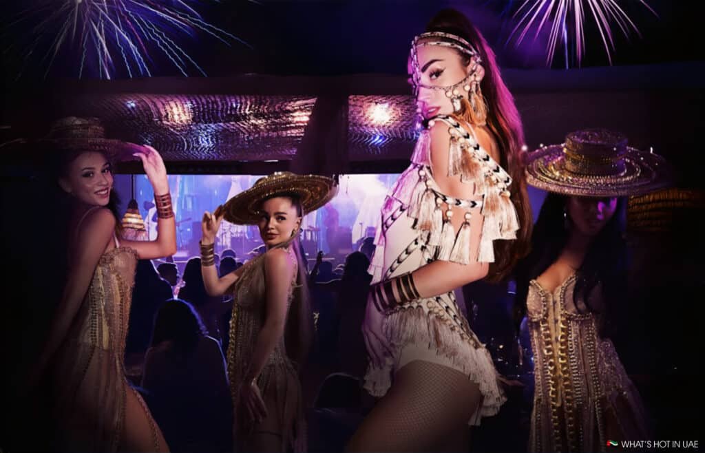 Four women in elaborate costumes and hats perform on Hedonïsm stage with fireworks in the background.