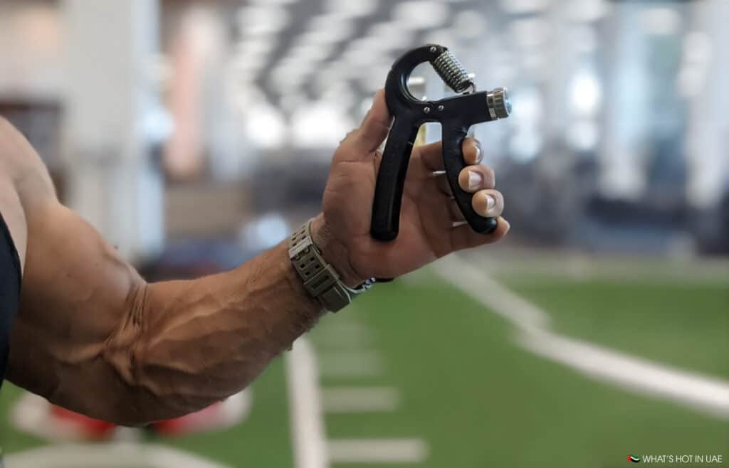 A person Improving Grip Strength holding a black hand grip strengthener in a gym setting.