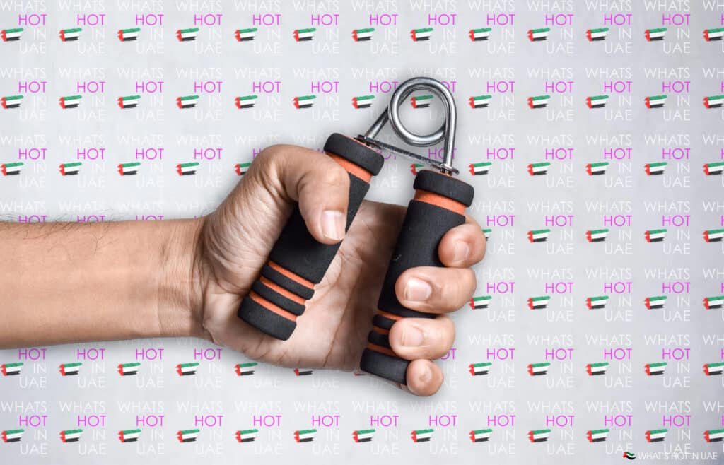 A hand holding a black and orange hand grip exerciser against a background with repeated text and UAE flags.
