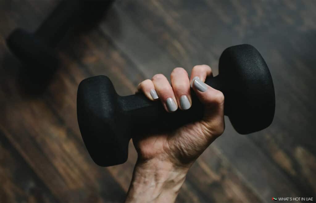 A hand with light gray nail polish holding a black dumbbell against a wooden floor background.