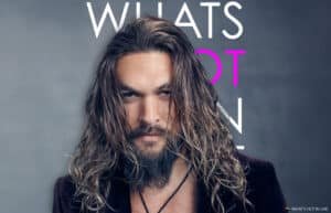 A man with long wavy hair and a beard poses against a background featuring partially visible text.