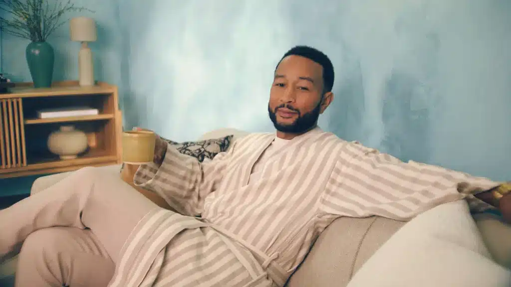 A man in a striped robe lounges on a sofa, holding a mug in a relaxed setting with a light blue wall.