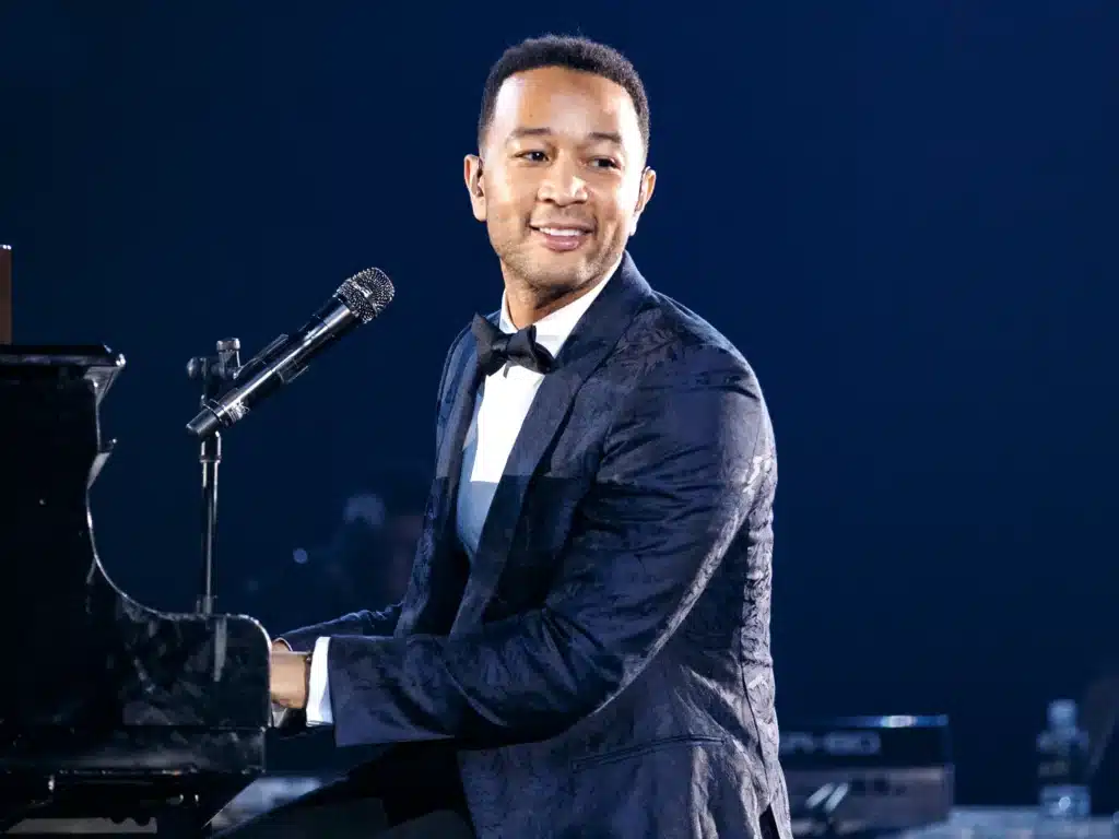 John Legend in a dark suit playing the piano and singing on stage, under bright lights.