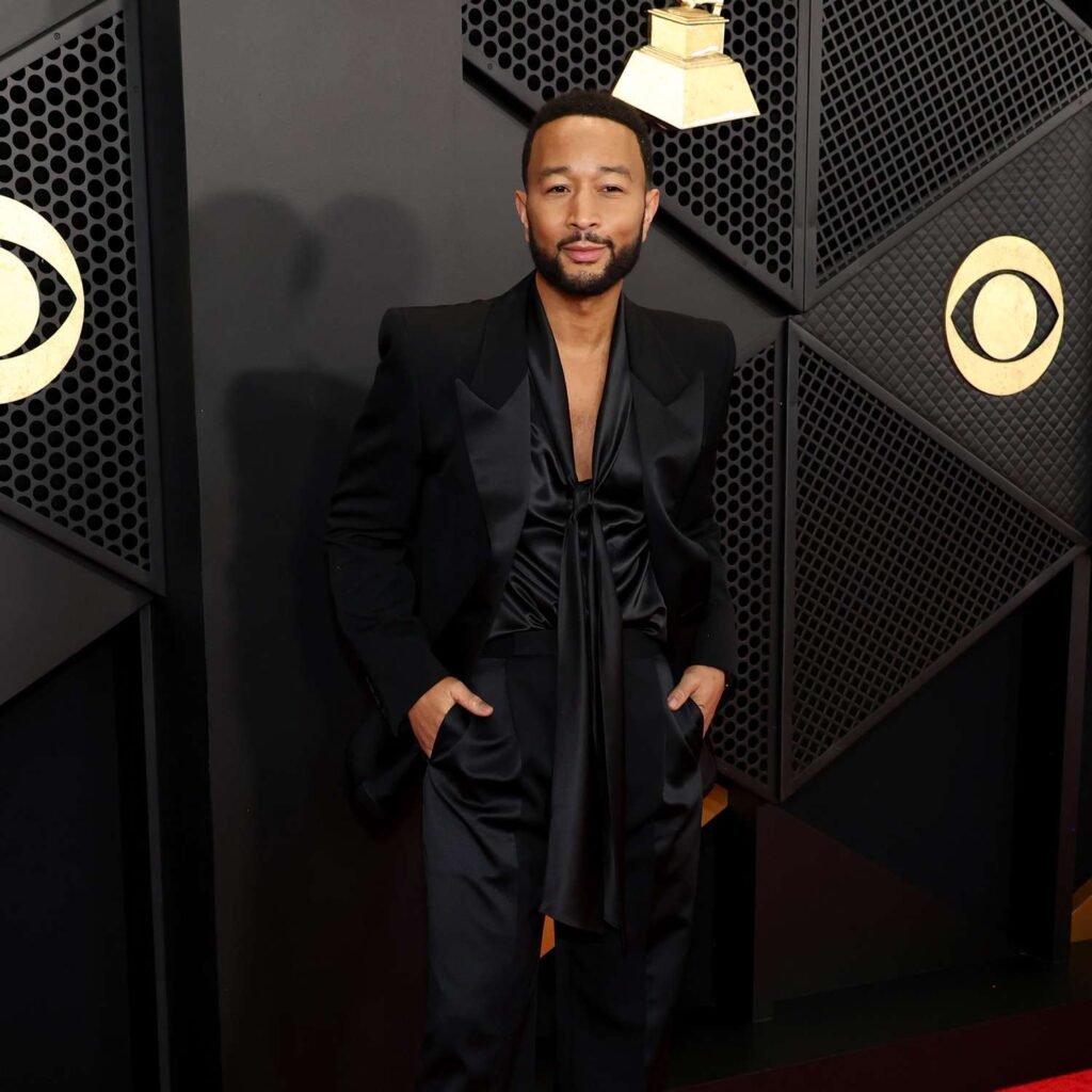 John Legend in a black suit with a silk shirt stands with hands in pockets against a geometric black wall with gold accents.